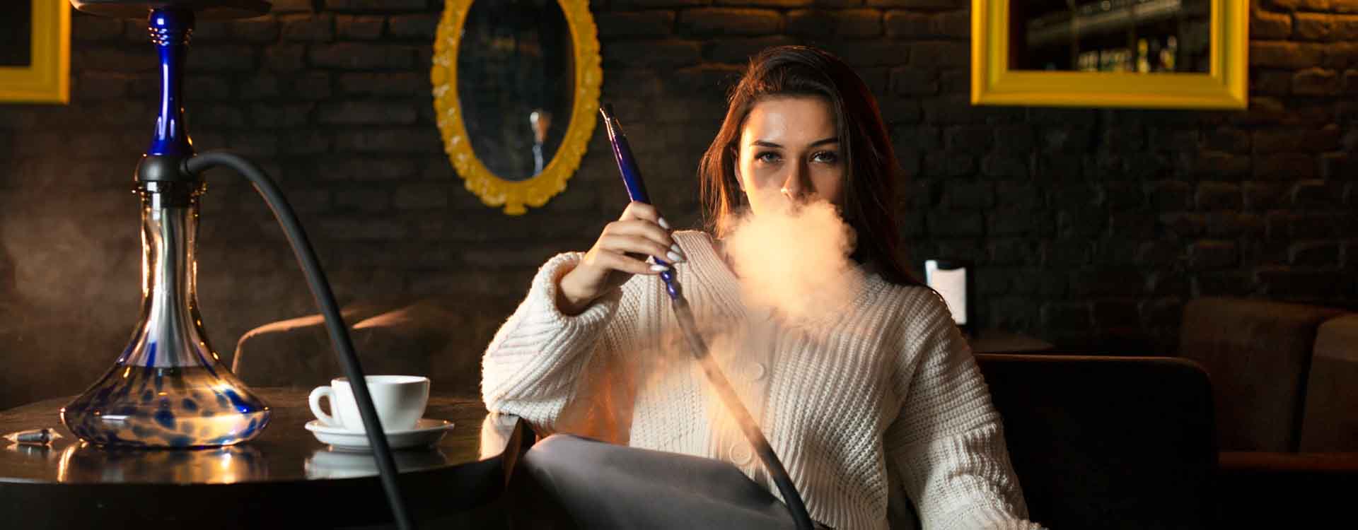 Mladá žena v shisha bare vydychuje dym z vodnej fajky a pozerá sa priamo do objektívu kamery